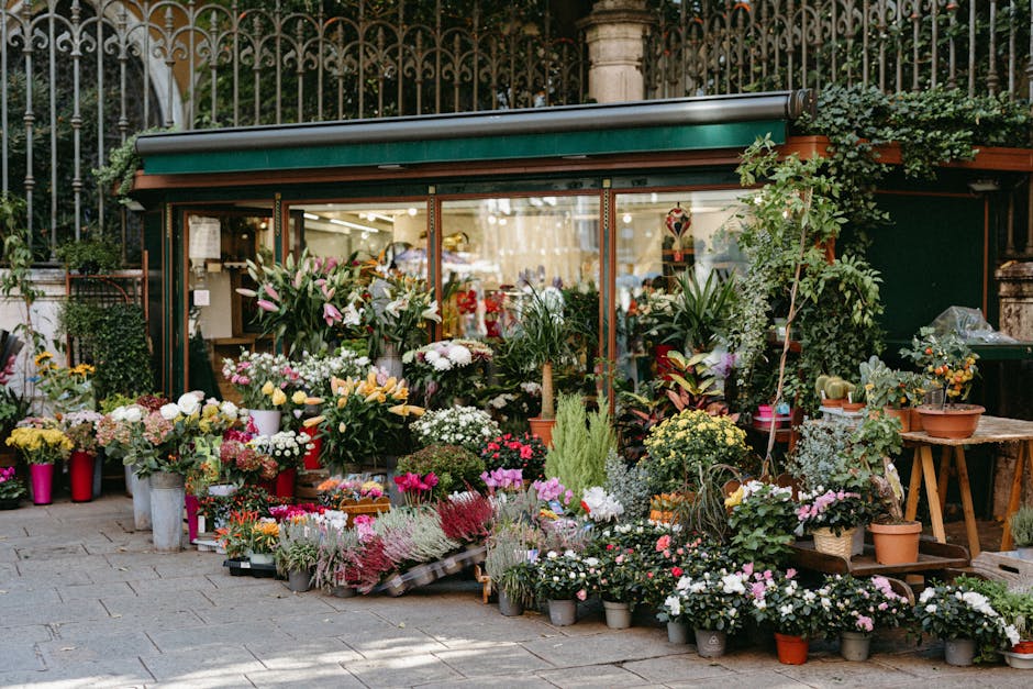 An outdoor flower shop with a green awning and ornate metal fencing behind, displaying a vibrant array of fresh flowers including roses, lilies, chrysanthemums, and foliage in various shades of pink, 