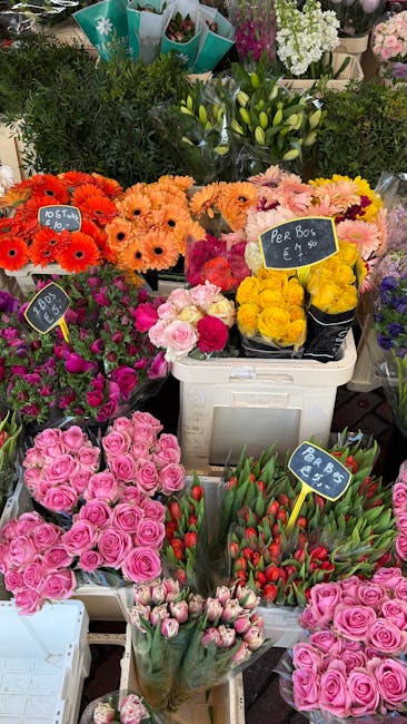 A vibrant display of fresh floral bouquets and potted flowers at a flower shop, featuring clusters of pink roses, red tulips, and yellow roses arranged in white crates and black buckets. The roses are